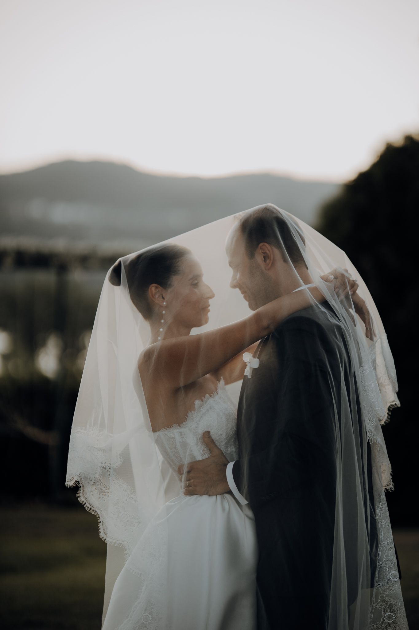 Bride and groom share romantic moment under wedding veil outdoors with mountain backdrop at dusk.