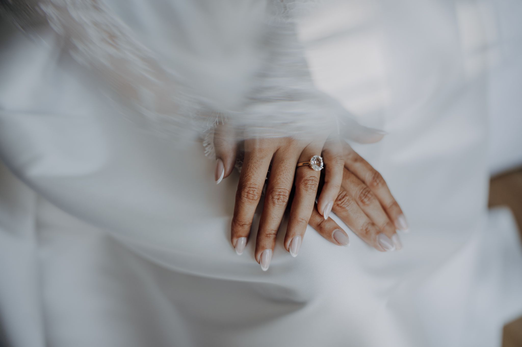 Close-up of bride's hands with white manicure and diamond engagement ring draped in flowing white wedding veil.