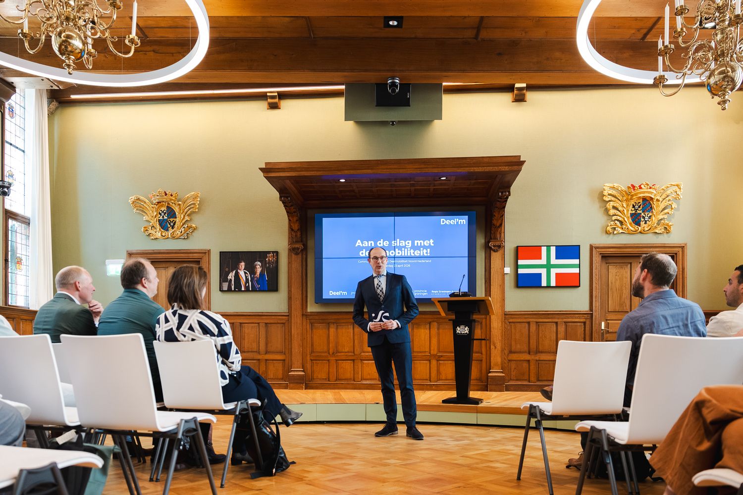 Professional speaker presenting at a formal conference in an elegant hall with coats of arms and seated audience.