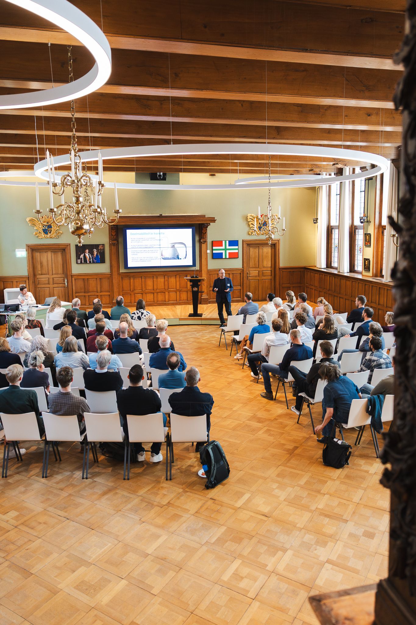 Audience seated in elegant wood-paneled conference hall listening to a presenter at a podium with projection screen.