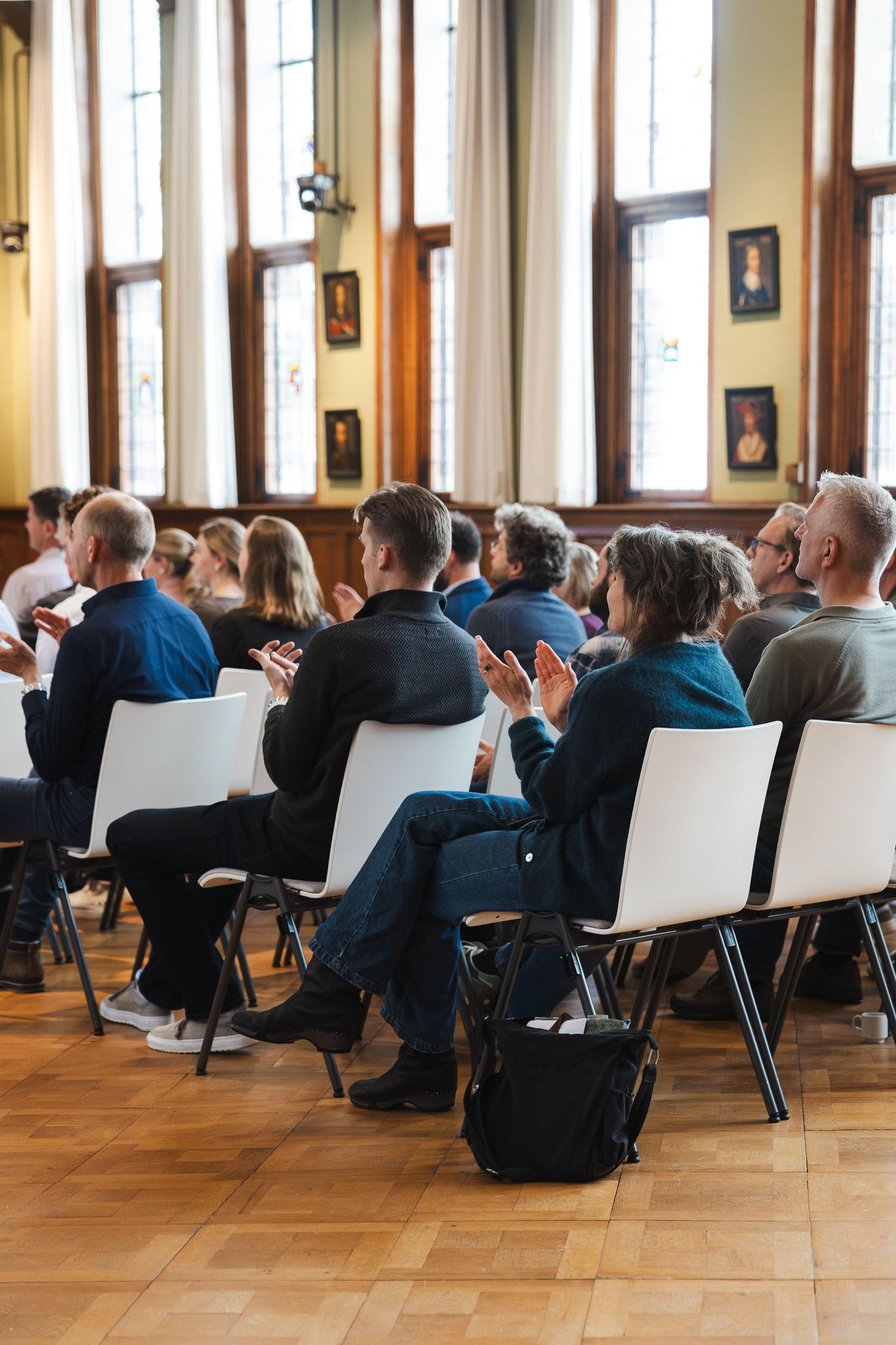 Audience members applauding in a elegant hall with wooden floors, white chairs, and portrait paintings on the walls.