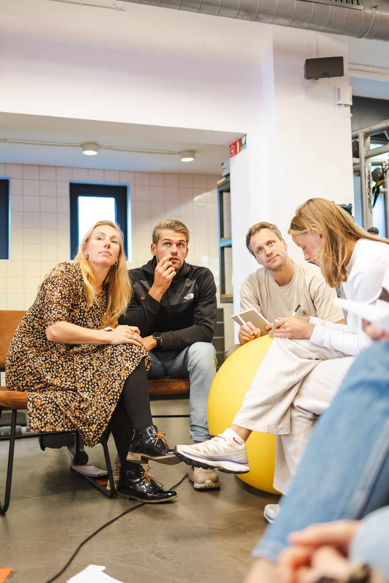 A group sits casually in conversation in a modern office space with white tiled walls and yellow exercise ball.