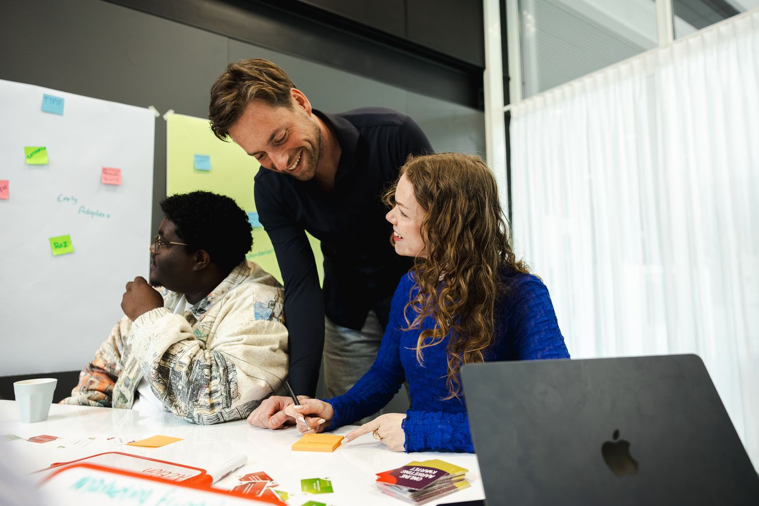 Three colleagues collaborate around a table with sticky notes and documents in a modern office workspace.