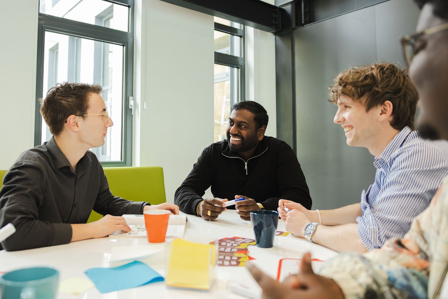 Diverse group of young professionals smiling and talking around a white office table with colorful cups and papers.