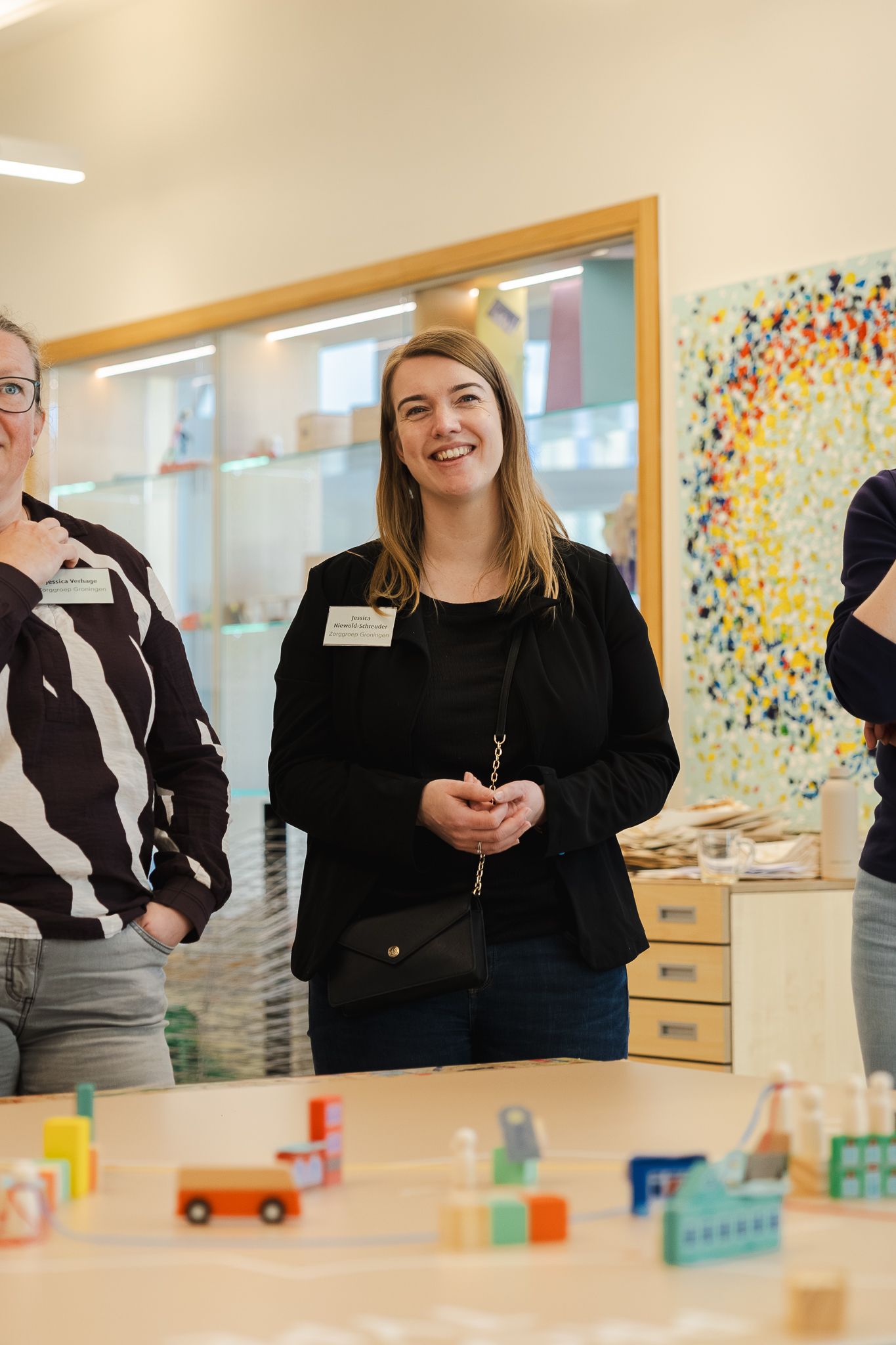 Three women wearing name badges stand smiling near a table with colorful wooden toys in a bright modern room.