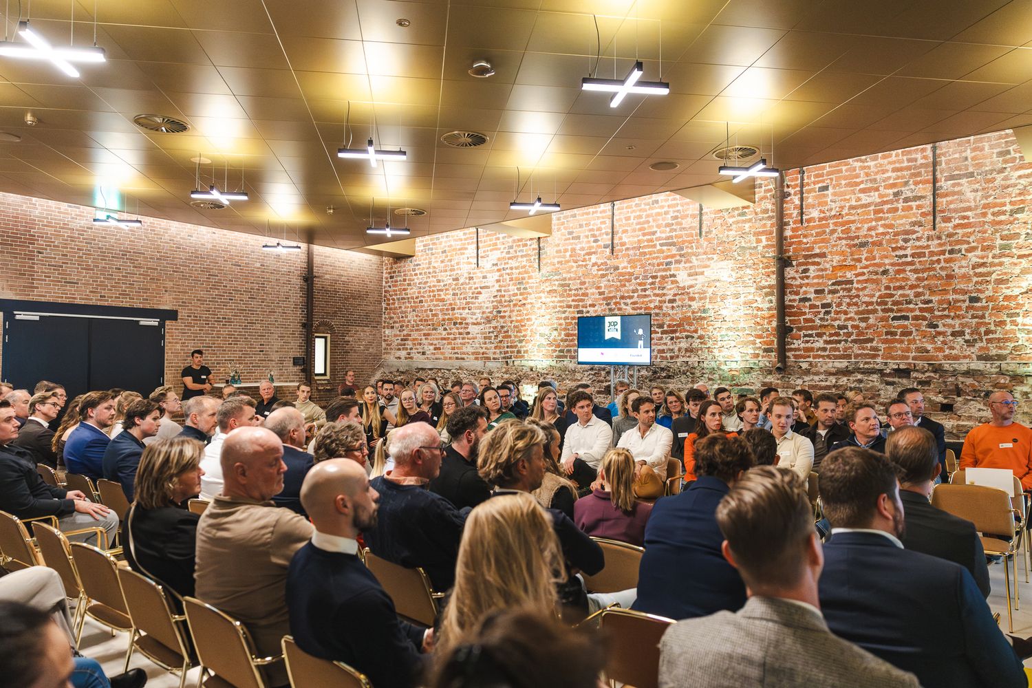 A crowded room with exposed brick walls and overhead lighting filled with people seated in rows for an event.