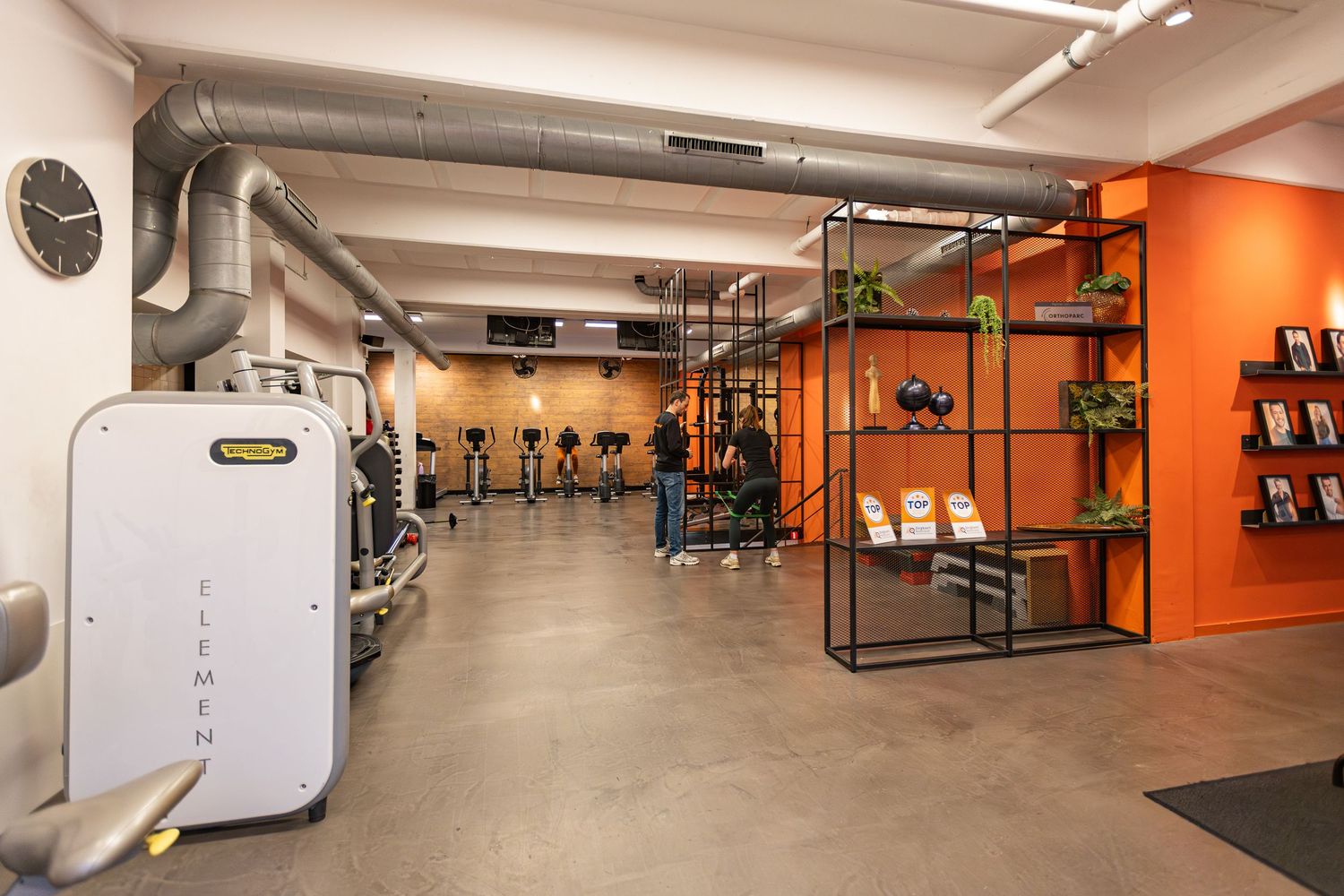 Modern fitness gym interior with orange accent wall, weight training equipment, and industrial style lighting fixtures.