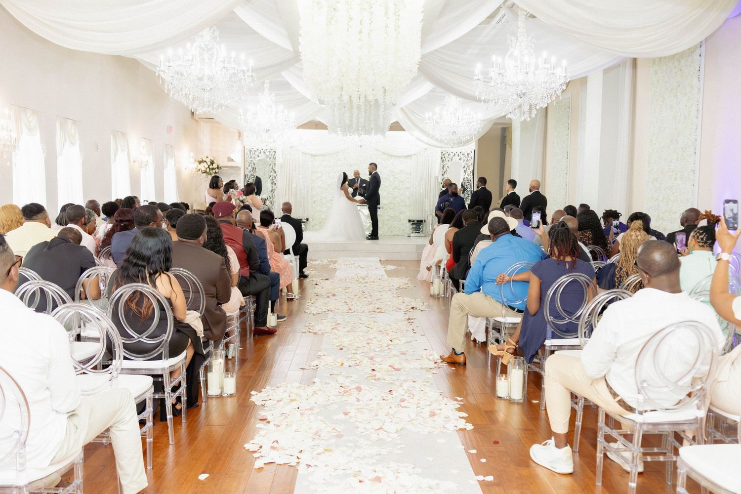Elegant all-white wedding ceremony with guests seated along a rose petal aisle under draped chandeliers.