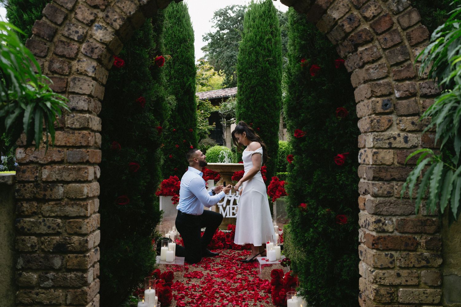 A romantic proposal moment under a brick archway decorated with red rose petals.