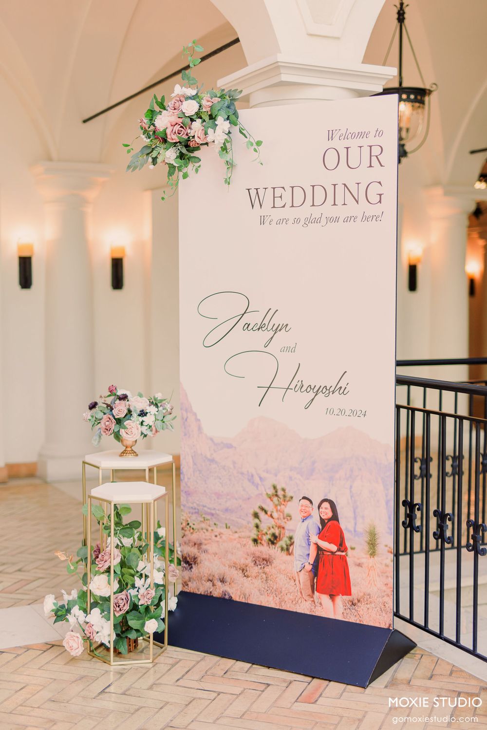 Elegant wedding welcome sign with floral decorations and desert mountain backdrop displayed in a white interior venue.