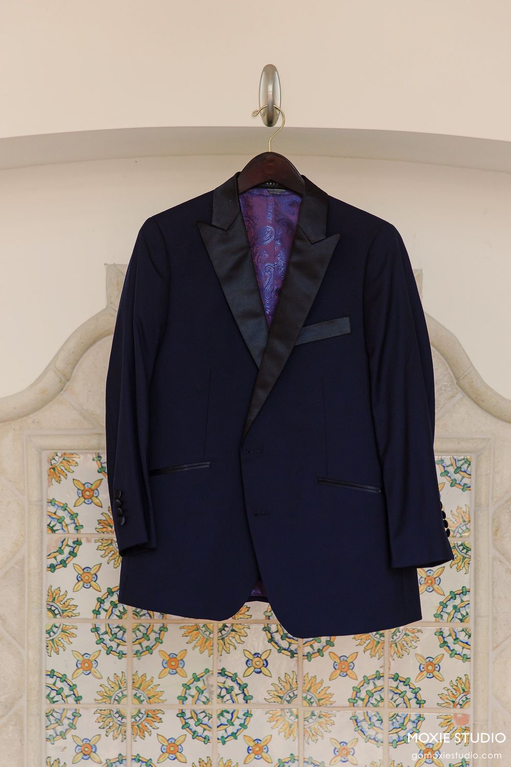 A navy blue tuxedo jacket with black satin lapels hangs on a white wall with floral wallpaper.