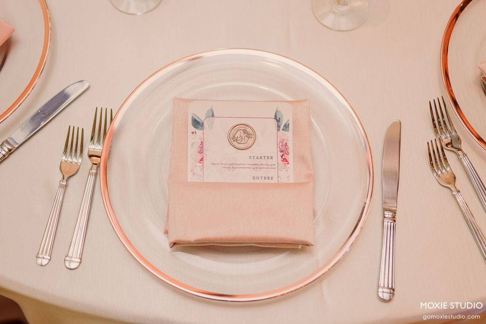 Modern place setting with rose gold charger plates, white dishes, and metallic flatware on a blush pink tablecloth.