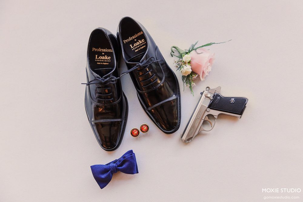 Black leather dress shoes with bow tie and floral accessories arranged on light beige surface for wedding photography.