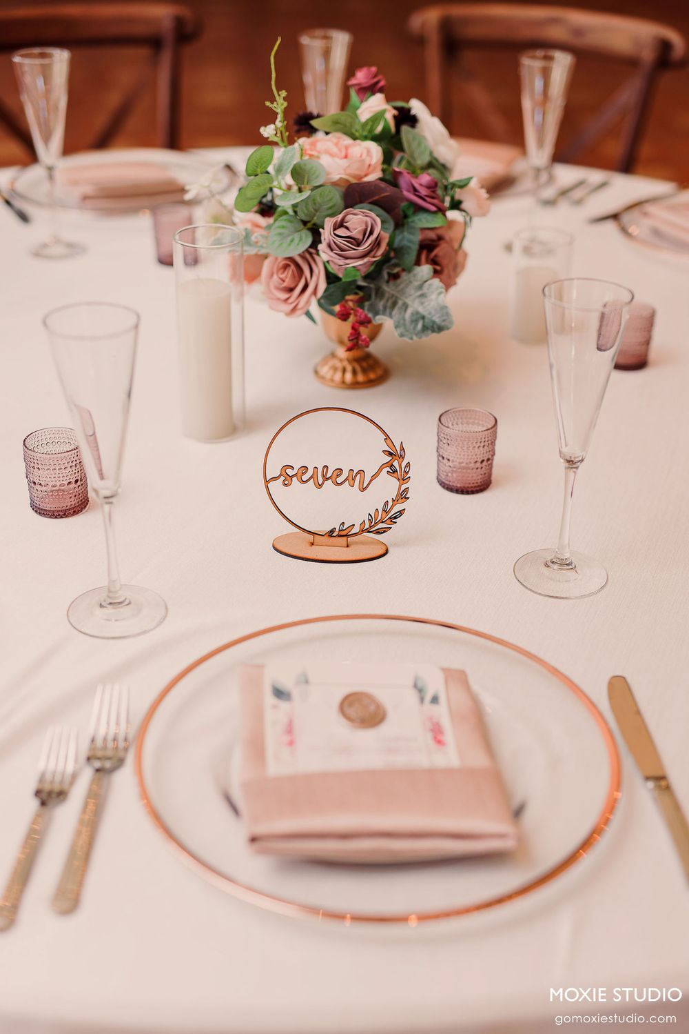Elegant wedding table setting with pink roses centerpiece, champagne glasses, and wooden table number in rose gold tones.