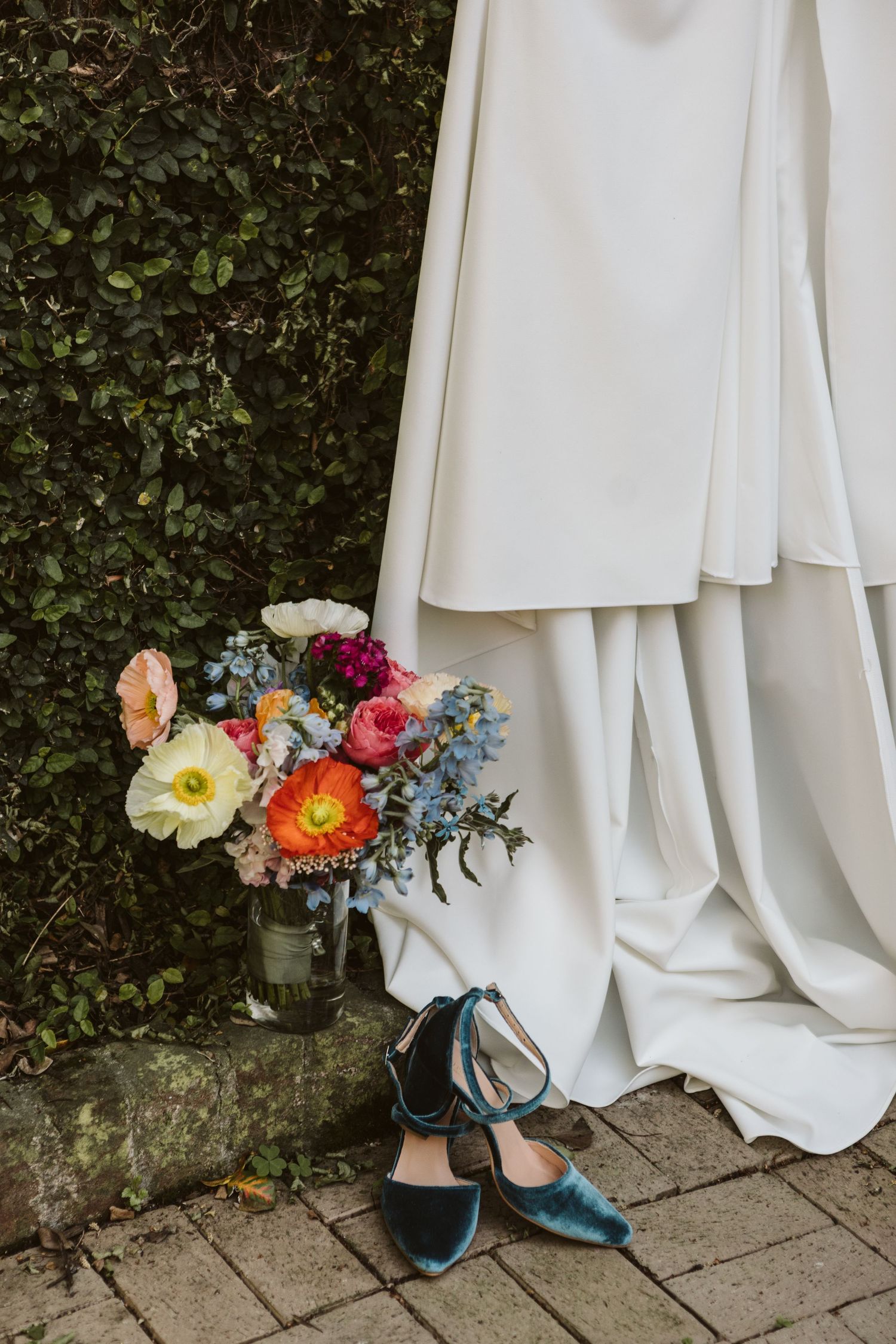 White wedding dress with colorful floral bouquet and teal shoes against garden wall.