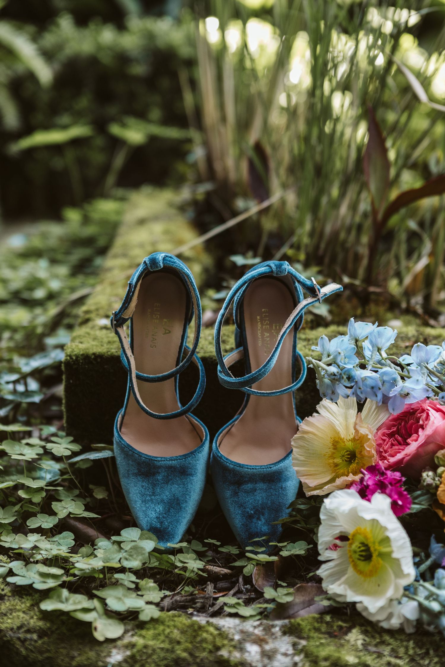 Vintage teal velvet wedding shoes surrounded by colorful flowers on mossy ground.