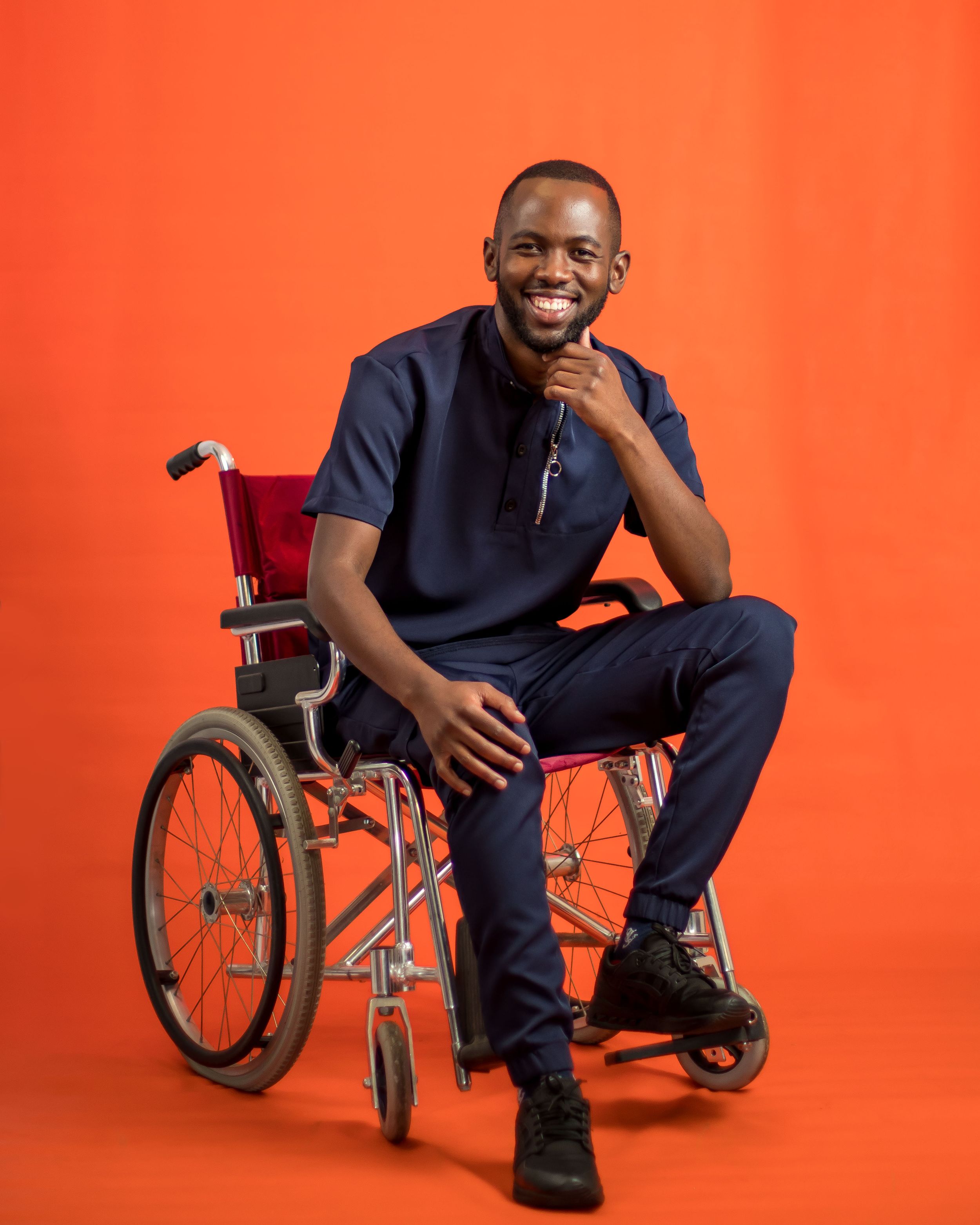 Seated in wheelchair wearing navy outfit against orange background.