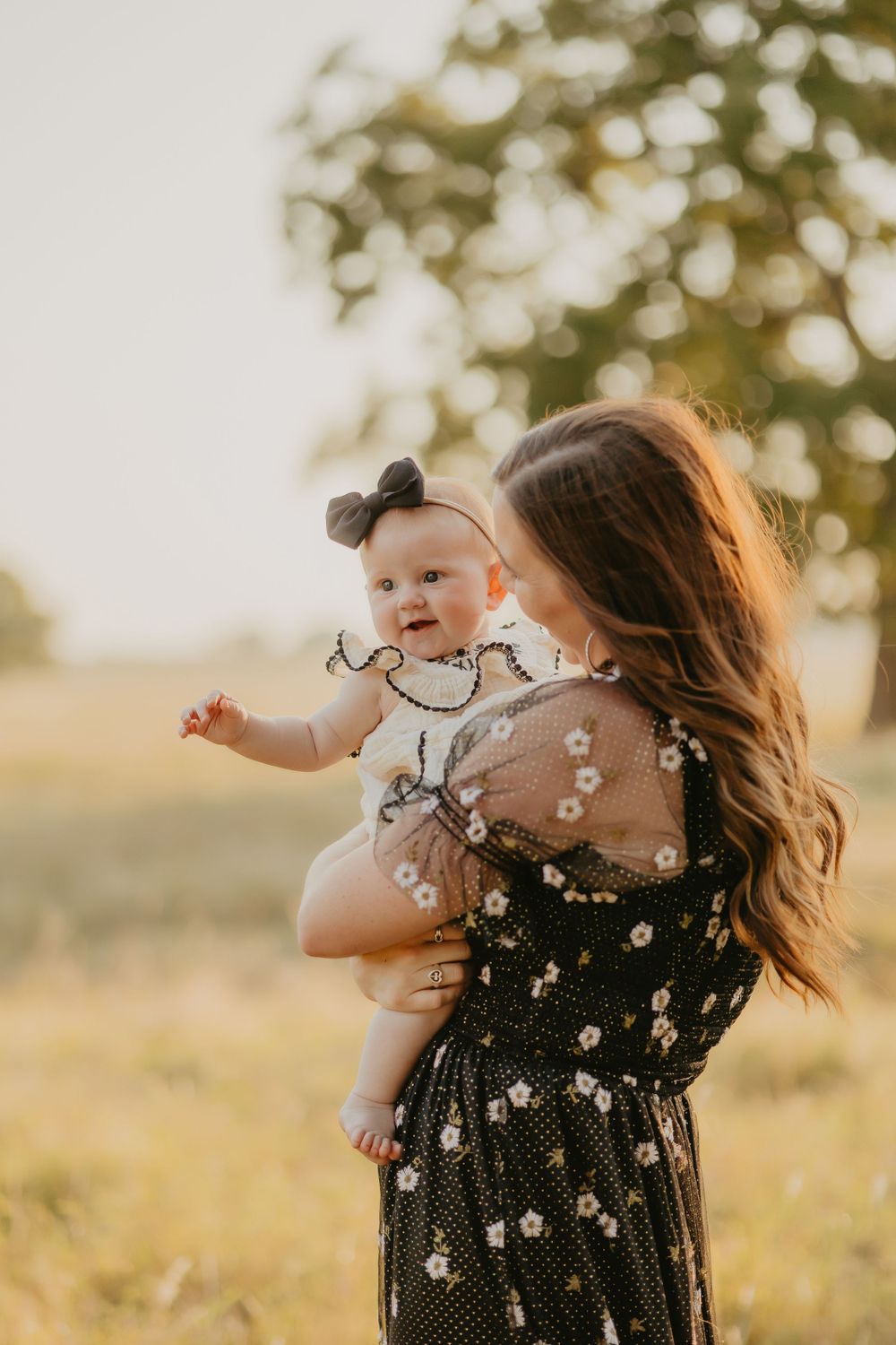 Mother and baby share a joyful moment in a floral dress against golden sunset light in a grassy field.