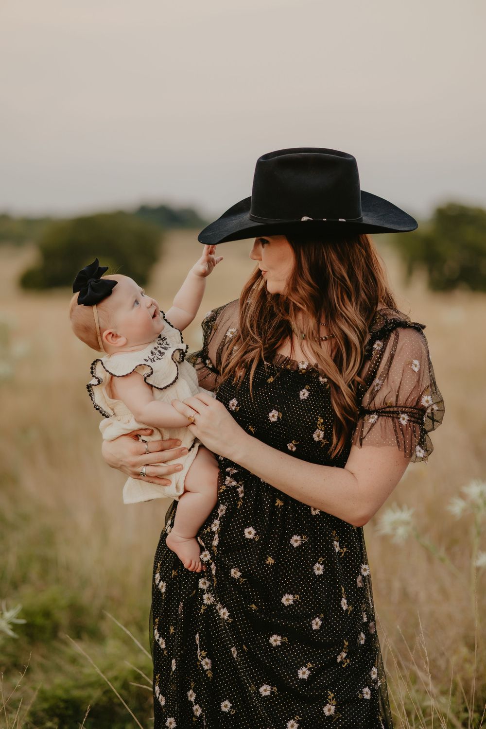 Woman in black floral dress and cowboy hat holds baby in rustic field setting.