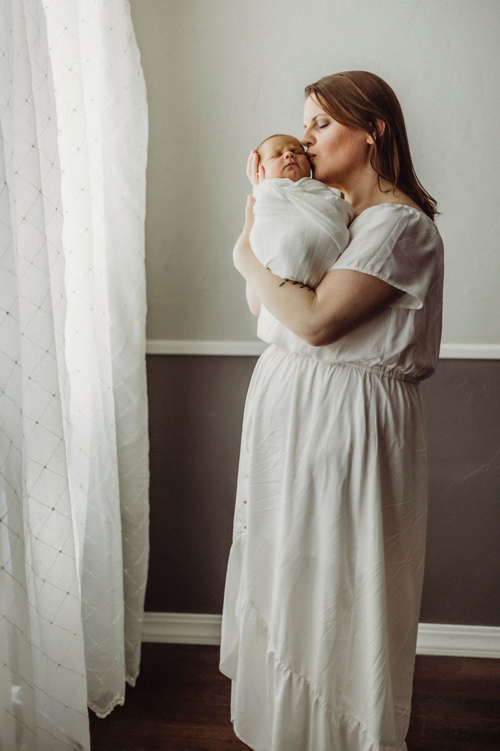 Woman in a flowing white dress holding a baby against a light wall.