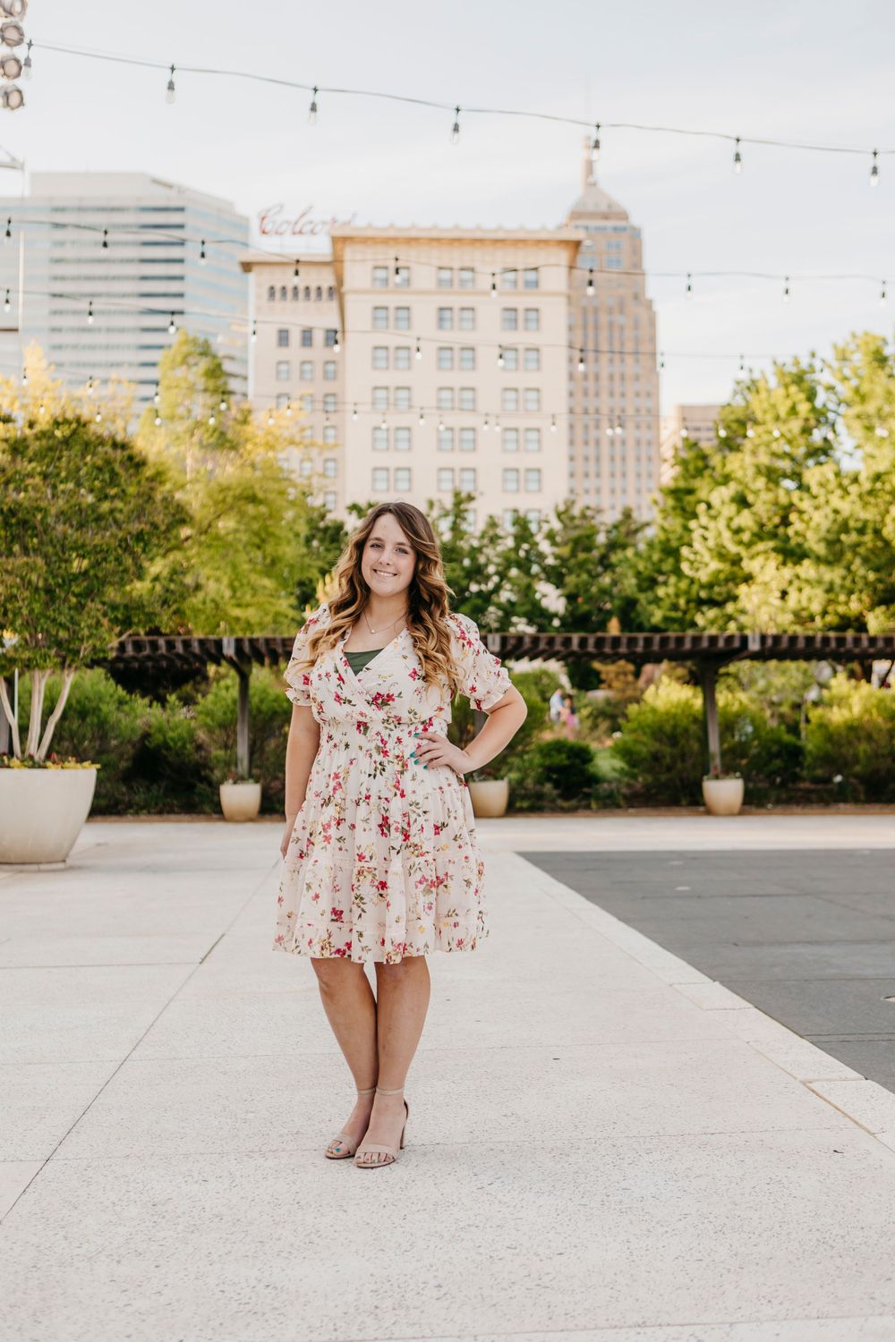 Woman in floral dress posing on rooftop terrace with city buildings in background.