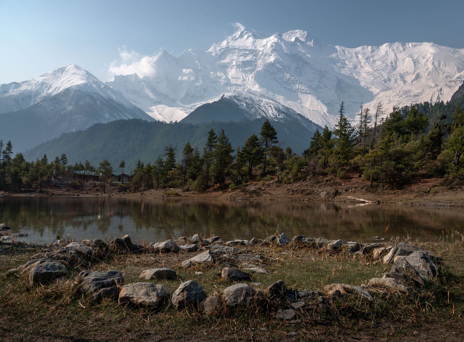 Alpine lake surrounded by rocks and pine trees with majestic Himalayan mountains in background.