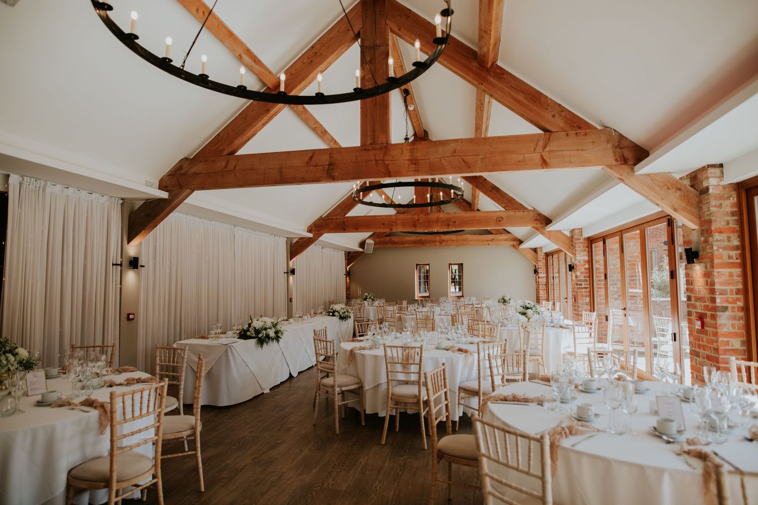 Reception tables with white linens set up in an elegant rustic barn venue with exposed wooden beams and natural lighting.