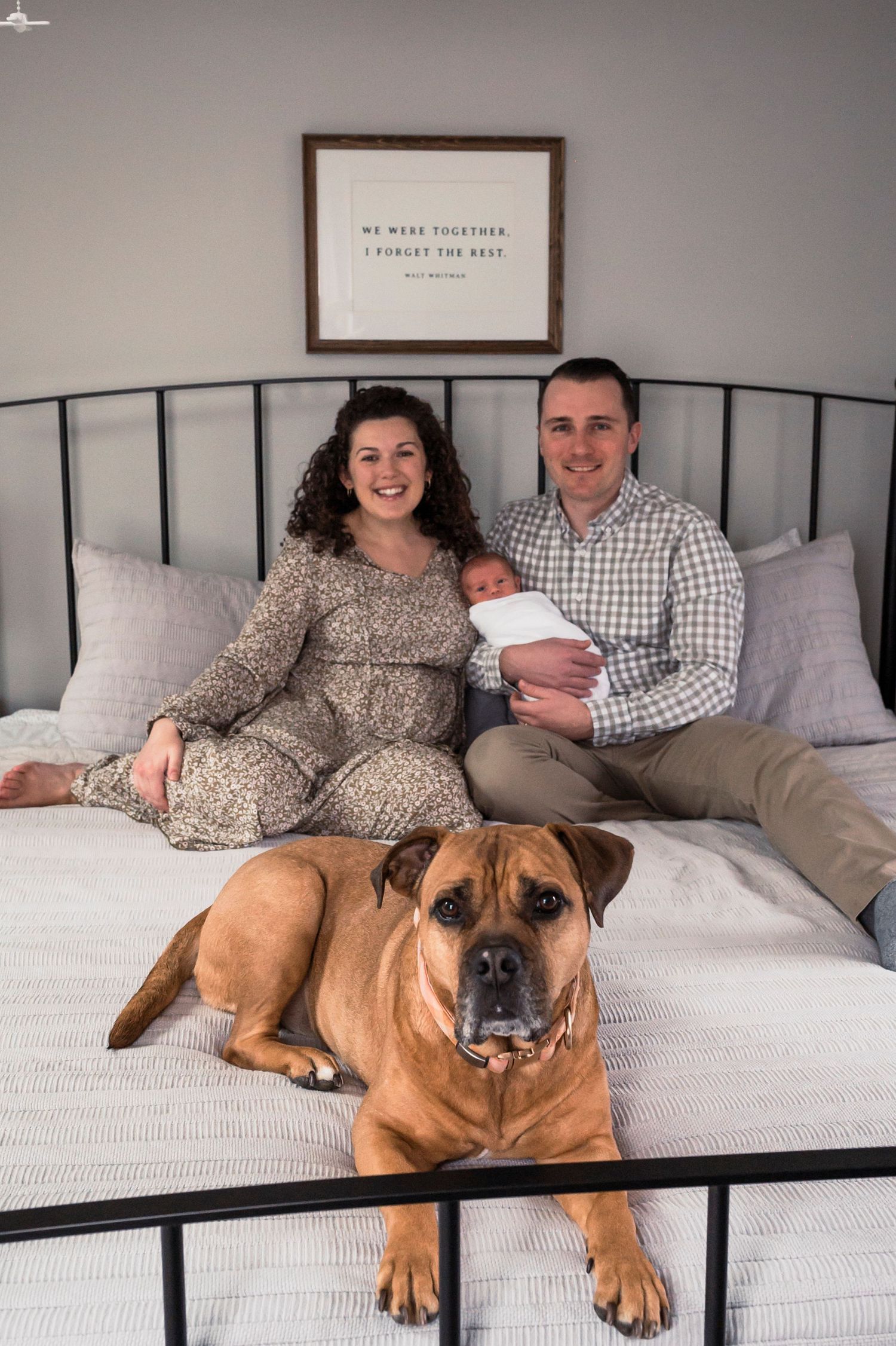 A family and their brown bulldog sit together on a bed beneath a framed wall quote in a cozy bedroom setting.
