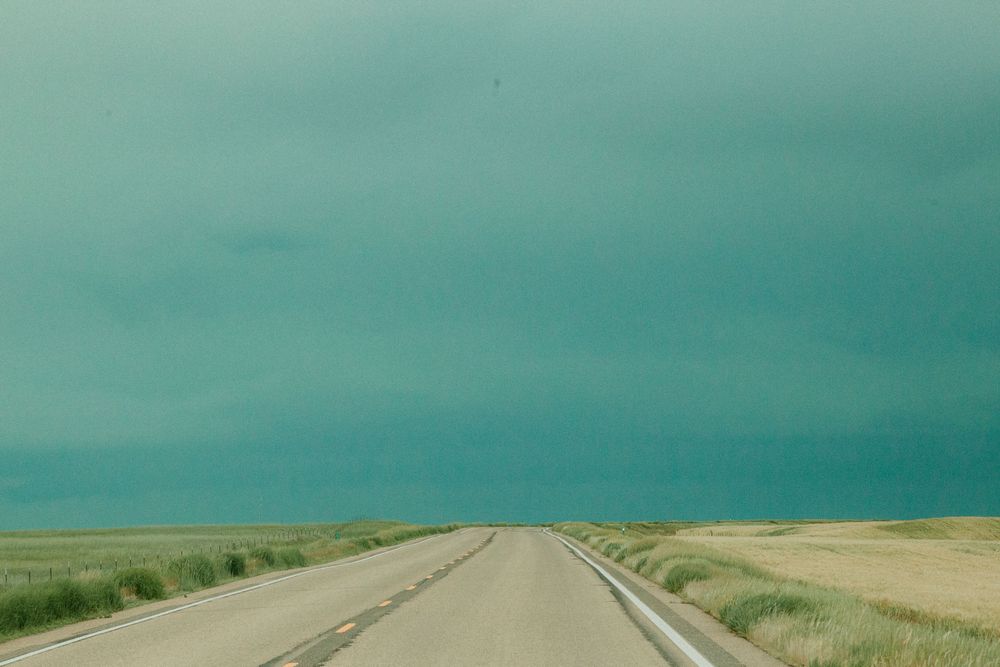 A flat, rural highway stretches toward the horizon under a moody turquoise sky in the Great Plains.