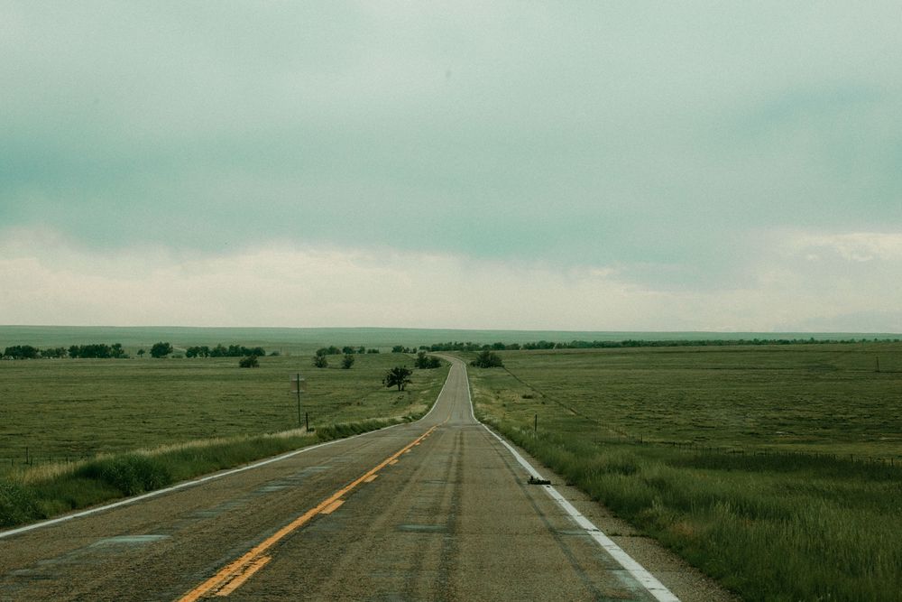 Long straight two-lane highway stretches into the horizon across flat grasslands under a cloudy turquoise sky.