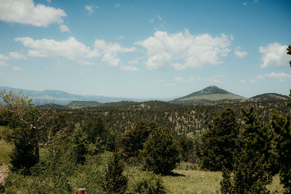 Scenic mountain landscape with rolling hills and evergreen forest under blue sky with white fluffy clouds.