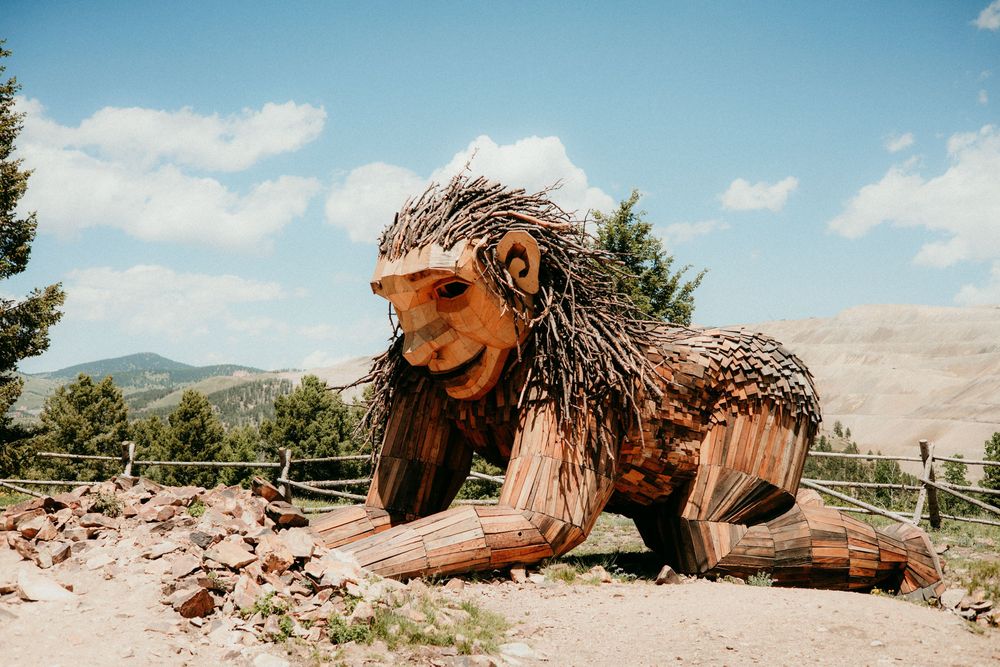 Large wooden lion sculpture rests on a hilltop with mountains and blue sky in the background.