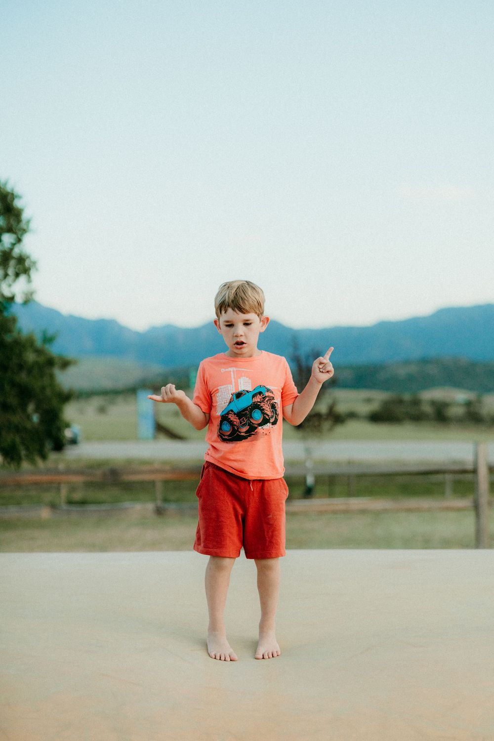 A young child in an orange shirt and red shorts stands barefoot outdoors with mountains visible in the background.