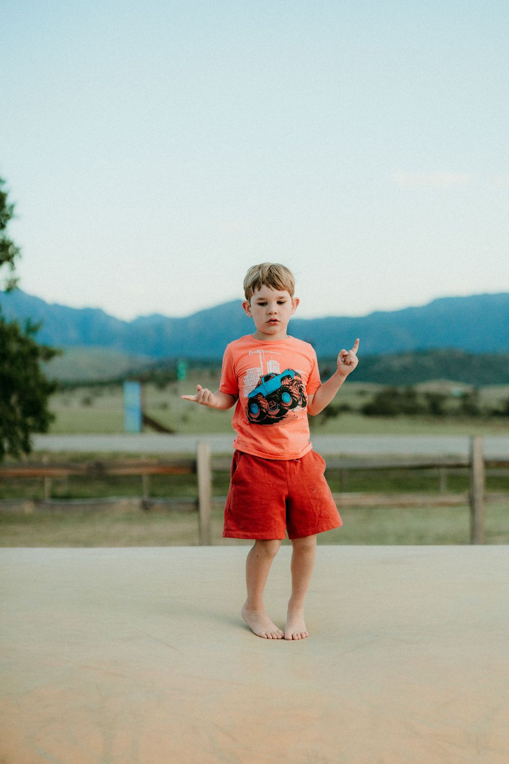 Small child in coral shirt and red shorts stands barefoot on concrete with mountains in background.