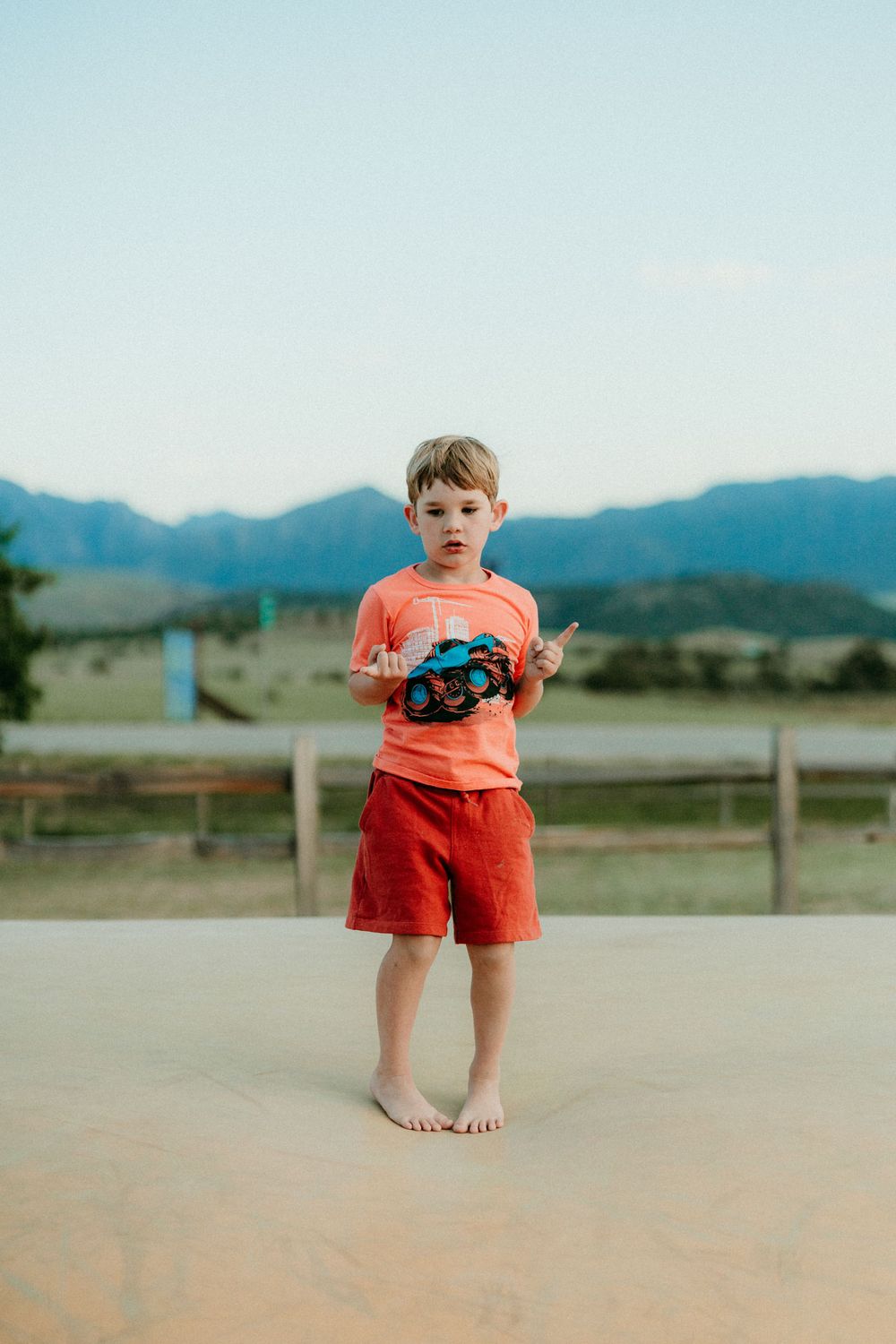 A barefoot child in orange clothing stands on an outdoor surface with mountains visible in the background at sunset.