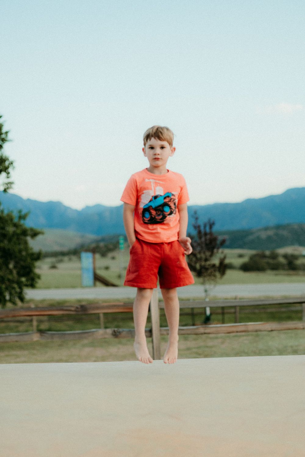 A figure in a bright coral shirt and shorts jumps on a path against a backdrop of rolling hills and mountains.