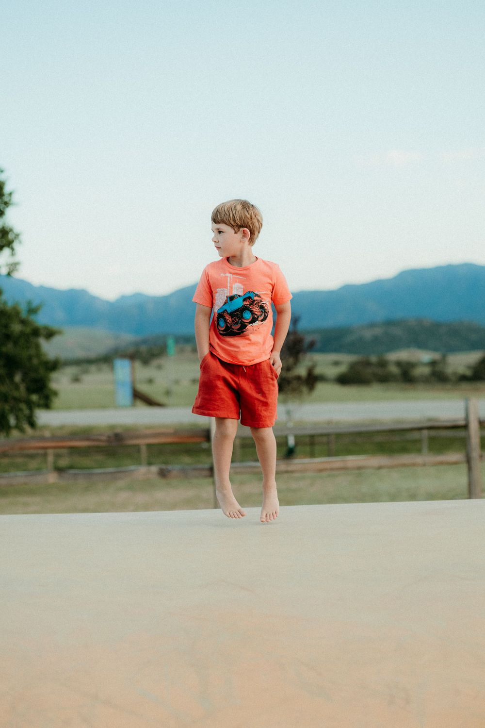 Four sequential photos show a small child in an orange t-shirt jumping up and down on a concrete surface with mountains behind.