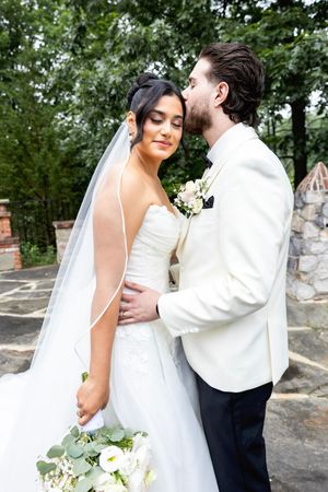 Elegant bride holds white floral bouquet while sharing tender moment with groom in white jacket outdoors. NJ wedding photographer