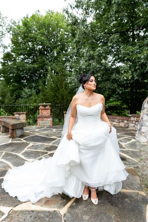 Beautiful bride in a strapless white wedding gown poses on a stone patio surrounded by lush green trees.