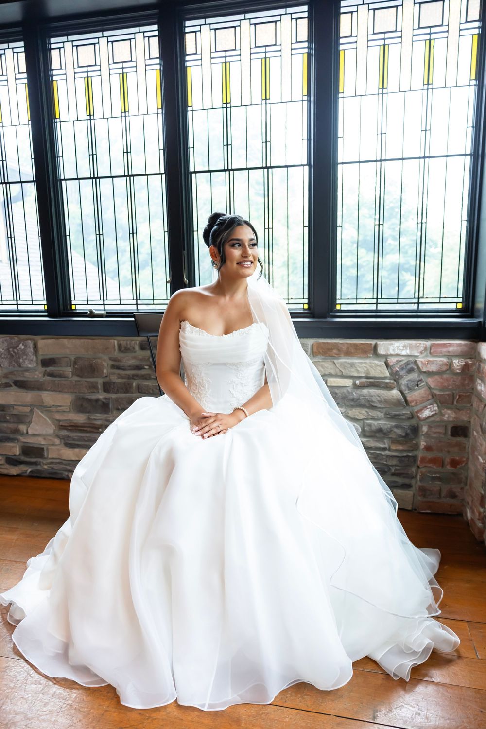 A bride in an elegant white strapless ball gown poses by a window in a venue with stone walls and hardwood floors. NJ wedding photographer
