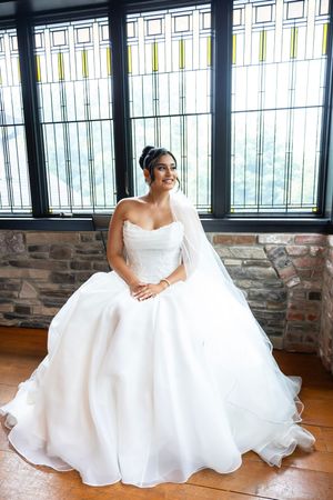 A bride in an elegant white strapless ball gown poses by a window in a venue with stone walls and hardwood floors. NJ wedding photographer