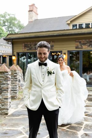 A wedding couple stands on a stone patio outside a rustic house with a chimney while wearing formal attire.