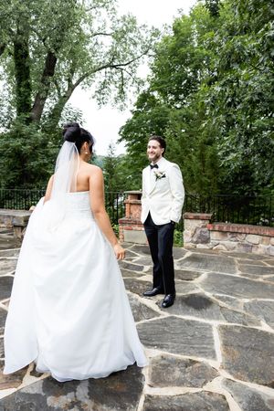 A couple during their first look moment standing on stone patio surrounded by lush green trees at an outdoor wedding venue.