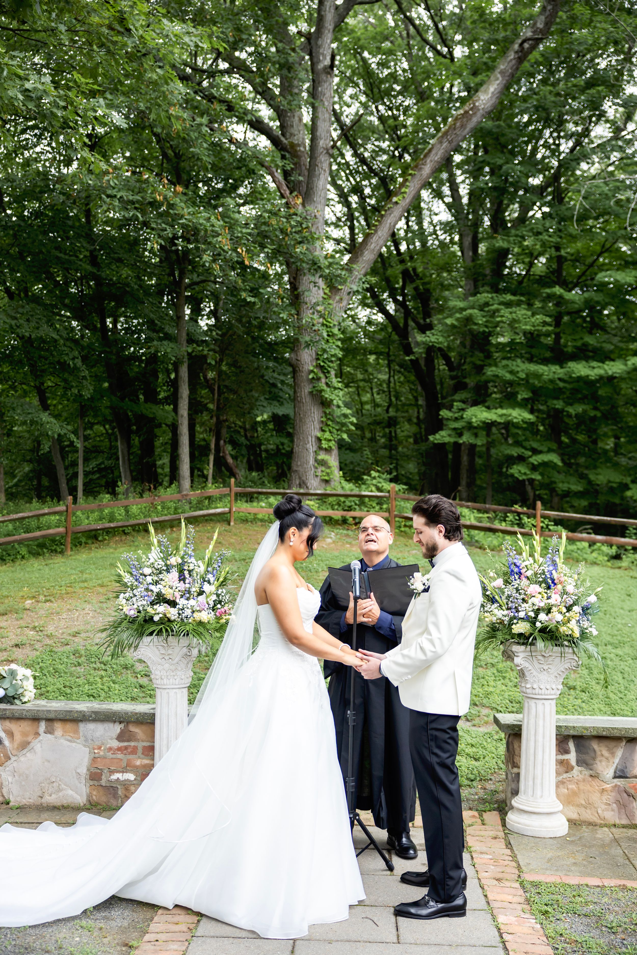 An outdoor wedding ceremony takes place in a wooded setting with stone patio and floral arrangements on white pedestals.