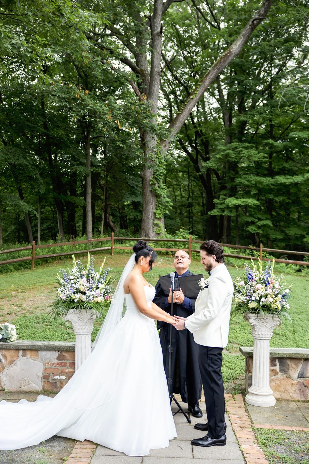 An outdoor wedding ceremony takes place in a wooded setting with stone patio and floral arrangements on white pedestals.
