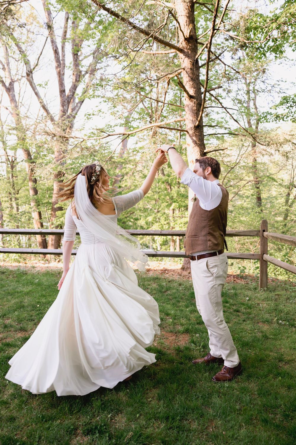 Bride and groom dance outdoors as groom twirls bride in white flowing wedding dress near wooden fence.