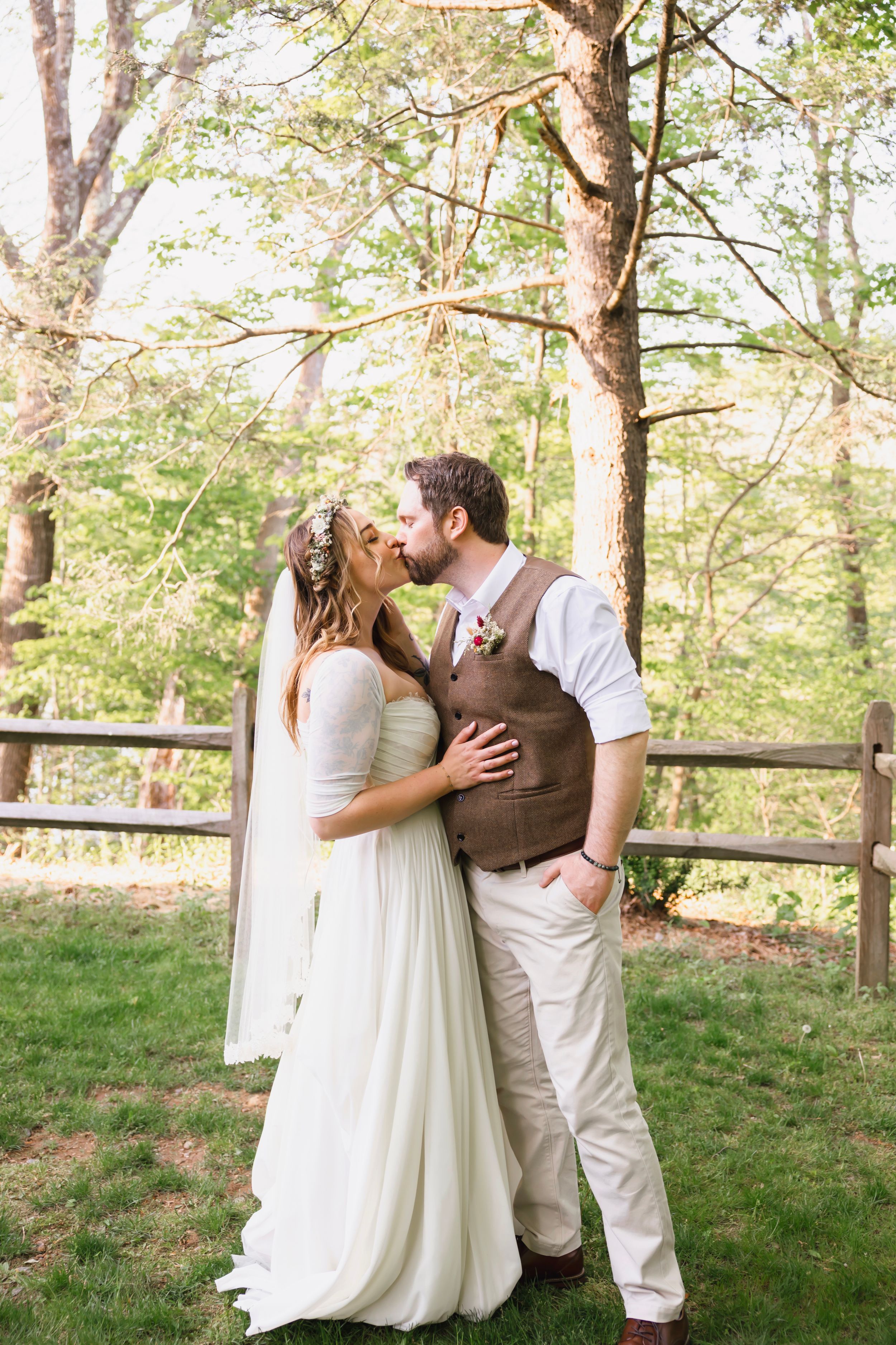 Bride and groom share a romantic kiss outdoors near a wooden fence surrounded by lush green trees on their wedding day.