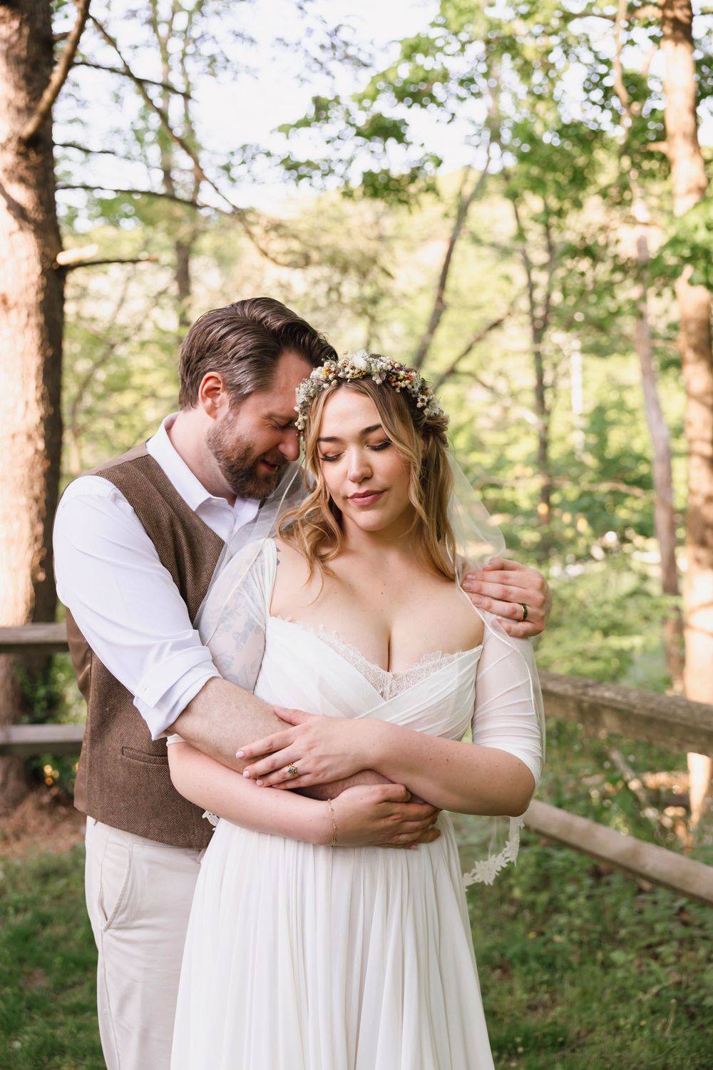 Groom in brown vest embraces bride wearing floral crown in white gown among sunlit forest trees.