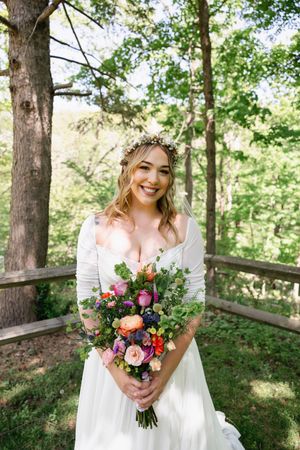 Smiling bride in white gown holds colorful bouquet wearing floral crown outdoors in lush green forest setting.