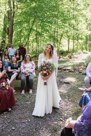 Bride walks down outdoor woodland aisle in white gown holding bouquet as guests watch the ceremony.